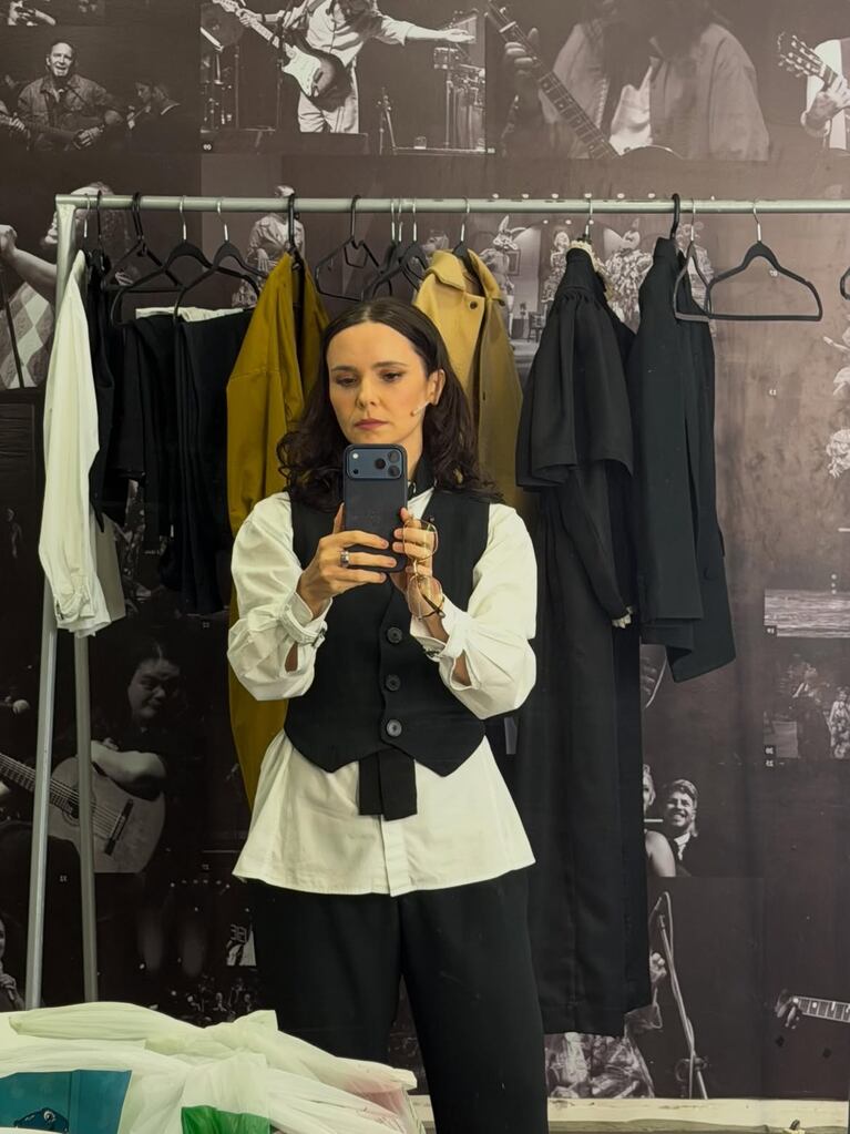 Débora Falabella tomándose una selfie frente a un espejo, vestida con chaleco negro y camisa blanca, rodeada de percheros con ropa de vestuario.