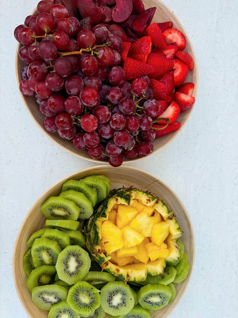 Dos bowls de madera con uvas, frutillas, kiwi y ananá sobre una superficie blanca.
Crédito Instagram.