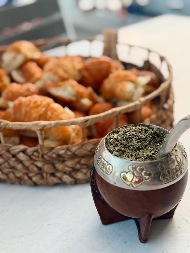 Mate de calabaza junto a una canasta con medialunas doradas sobre una mesa.
Crédito Instagram.